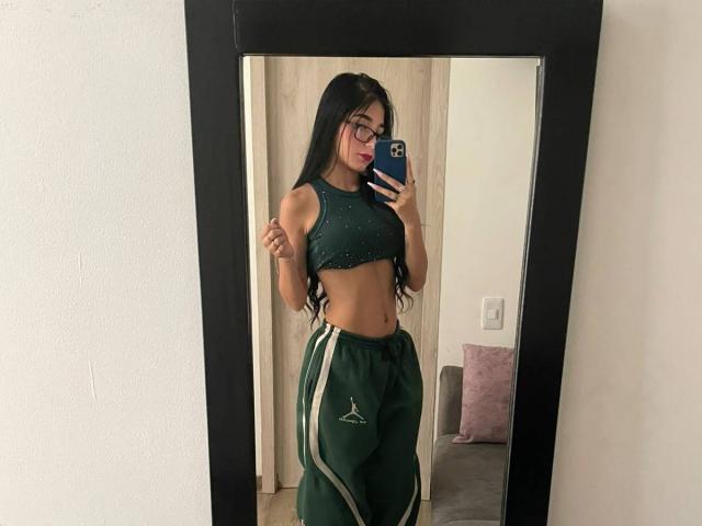 VioletaVelvet with long black hair, wearing a green sports bra and green track pants with white stripes, taking a mirror selfie, showing her toned midriff and small ass.