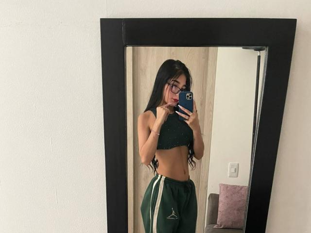 VioletaVelvet with long black hair, glasses, green crop top, showing her toned midriff and green track pants with white stripes, taking a mirror selfie.