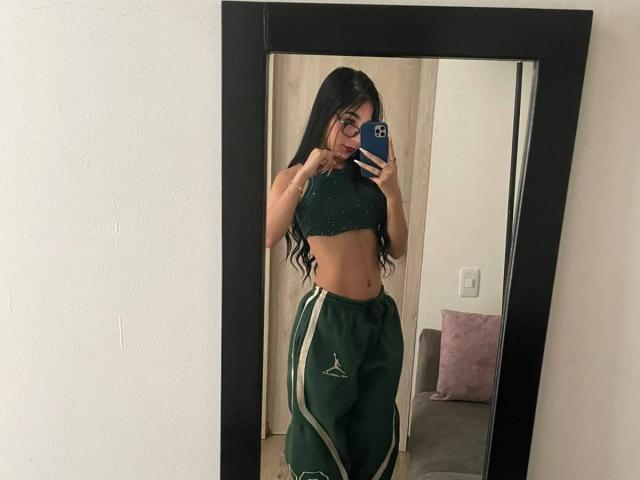 VioletaVelvet with long black hair, wearing a green crop top and green track pants with white stripes, taking a mirror selfie, showing her toned midriff and small ass.