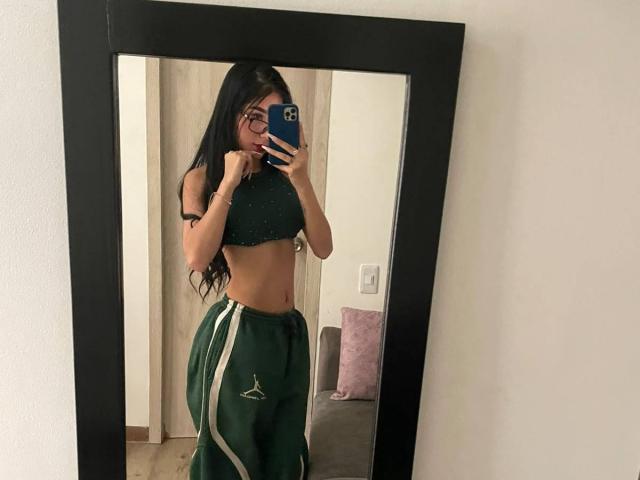 VioletaVelvet with long black hair, wearing glasses, green crop top, and green track pants with white stripes, taking a mirror selfie showing her toned midriff and ass.