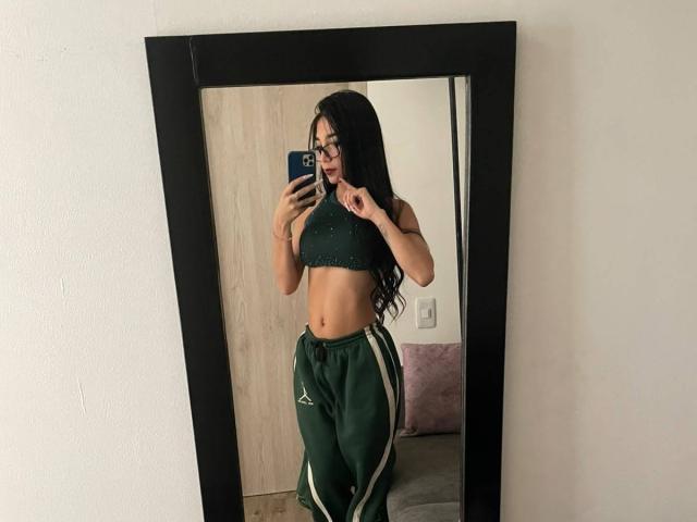 VioletaVelvet with long black hair, glasses, green sports bra, showing her toned abs, and green track pants with white stripes, taking a mirror selfie.