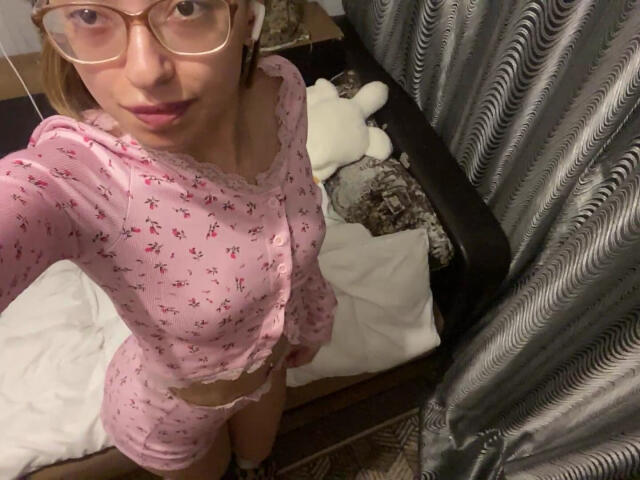 RaneeDerienzo mit Brille, in rosa, blumigem Pyjama, mit kleinen Brüsten und freiem Bauch, steht in einem Raum, blickt nach oben zur Kamera, im Hintergrund sind ein Bett und ein Stofftier zu sehen..