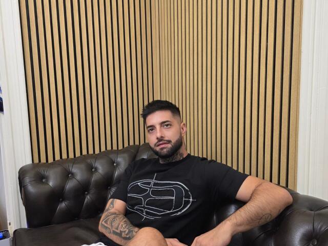 TommyDum, un hombre con barba y tatuajes, está sentado en un sofá de cuero marrón, vistiendo una camiseta negra con un diseño geométrico blanco, y mirando a la cámara..
