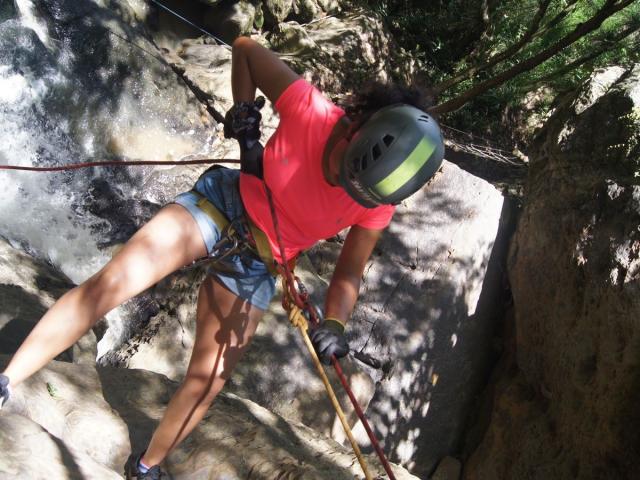KirasPell with curly hair, pink shirt, and denim shorts, climbing rock using ropes, wearing a green helmet, black gloves, and yellow harness, legs spread, hands gripping ropes.