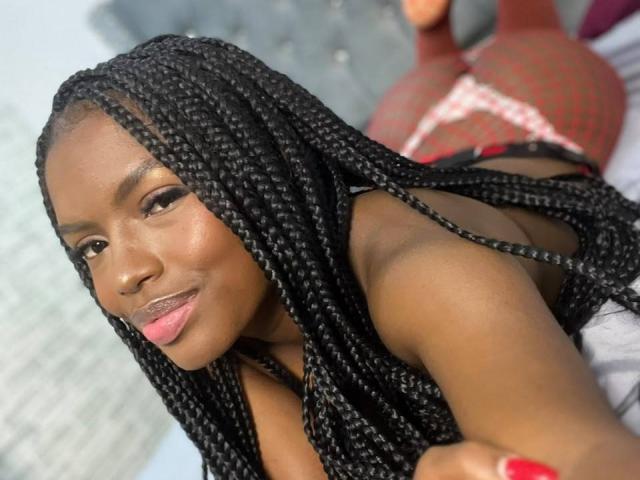 EllysRose, com pele escura, longas tranças pretas e batom rosa, deitada de bruços, vestindo lingerie preta, mostrando suas costas nuas e meias de rede vermelhas, sorrindo para a câmera..