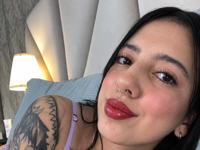 Hannah Cruz ha i capelli scuri, un piercing al naso, labbra rosse lucide e un tatuaggio sulla spalla. Indossa un reggiseno viola e guarda la fotocamera con un leggero sorriso..