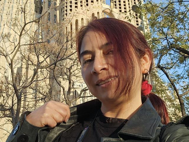 Alessia, capelli rossi, giacca nera e elastico per capelli rosso, tiene il colletto della giacca, in piedi di fronte a un edificio alto, beige, simile a un castello, sorride mostrando i denti..