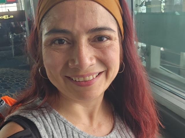 AlessiaRed hat rote Haare, einen braunen Stirnband und lächelt, wobei ihre kleinen Brüste sichtbar sind. Sie trägt ein graues Hemd und trägt Ohrringe im Hula-Hoop-Stil. Sie steht in der Nähe eines Glasfensters..