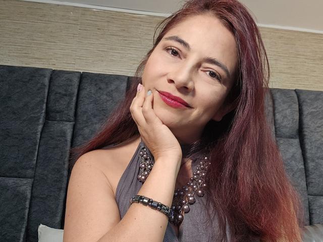 Alessia: capelli rossi, top grigio senza maniche, collana e braccialetto con perline, mano sul viso, sorridente, rossetto rosso e testiera grigia sullo sfondo..