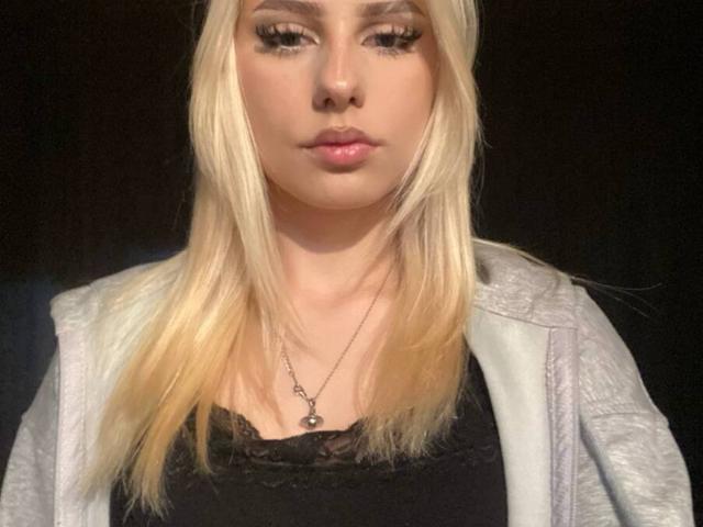 Blond JaneenOsher with long hair, wearing a black lace top, grey jacket, and necklace, has full lips and dark eyelashes, looking straight at the camera.
