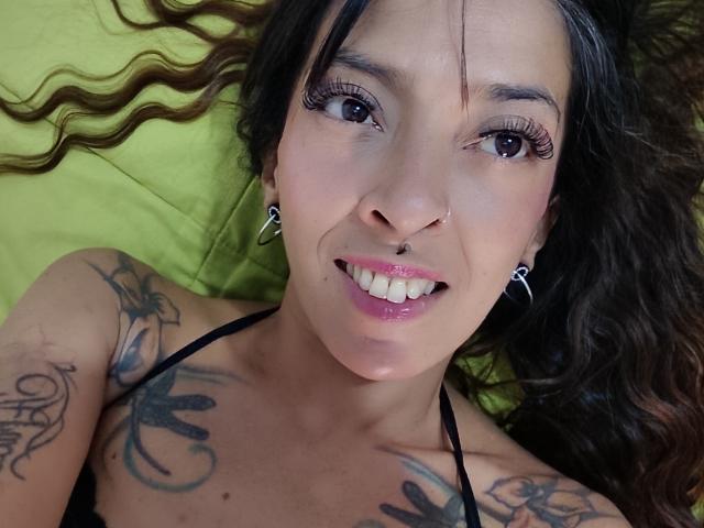 IsaChaud with dark wavy hair, nose ring, and tattoos on her chest and shoulder, smiling with pink lips, wearing black straps, and hoop earrings, lying on a green pillow.