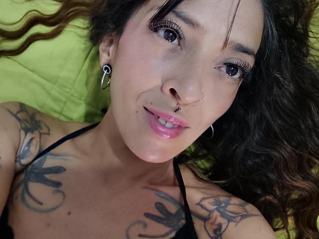 IsaChaud has curly brown hair, a nose ring, and a lip ring. She has tattoos on her chest and wears black eyeliner with long lashes. She's lying on a green pillow.