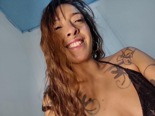 IsaChaud smiling with wavy brown hair, nose ring, hoop earrings, black lace top, tattoos on chest and shoulder, showing tits, with pink lipstick, in a selfie.