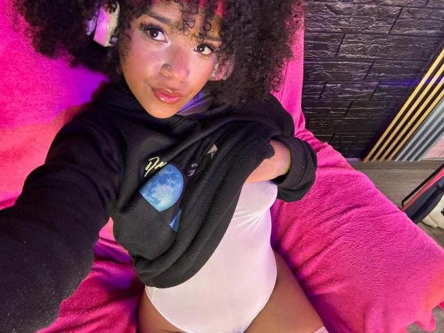 SerenaSiren with curly hair, pink lipstick, and nose ring, lifting black hoodie to reveal white one-piece, showing her small, round tits, and pink blanket. She's sitting, taking a selfie.