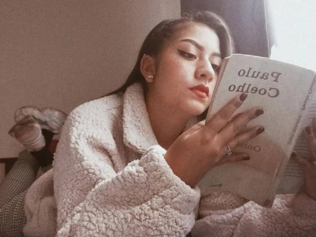 PaulaLoughty, with puffy white fleece coat, reading book, has full lips, dark brown hair, and gold earrings, showing her ring and painted nails. Soft light, relaxed vibe.