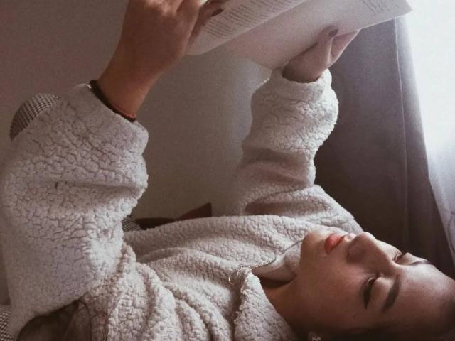 PaulaLoughty is lying on her back, reading a book, wearing a fluffy white sweater, with her legs up, looking relaxed, and has red lipstick.