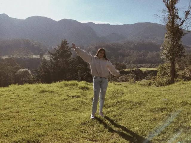 PaulaLoughty stands on a grassy hill, arms raised, wearing a white blouse and blue jeans, with a tree on the right and mountains in the background.