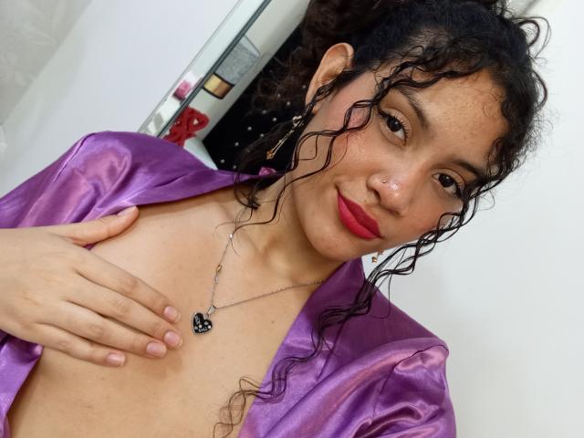 ValeryPoleth with curly black hair, purple satin robe, red lipstick, hand on chest, necklace with black anchor pendant, smiling, light brown skin, small breasts, white background.