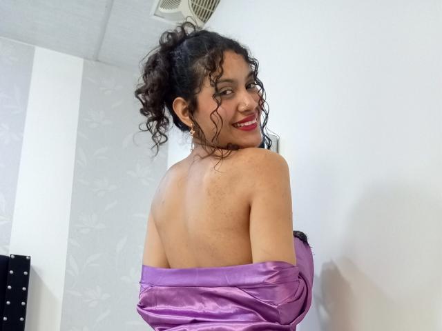 ValeryPoleth with curly black hair, smiling, back to camera, purple satin off-shoulder top, exposing her back and a hint of her bare ass. Red lipstick, white wall.
