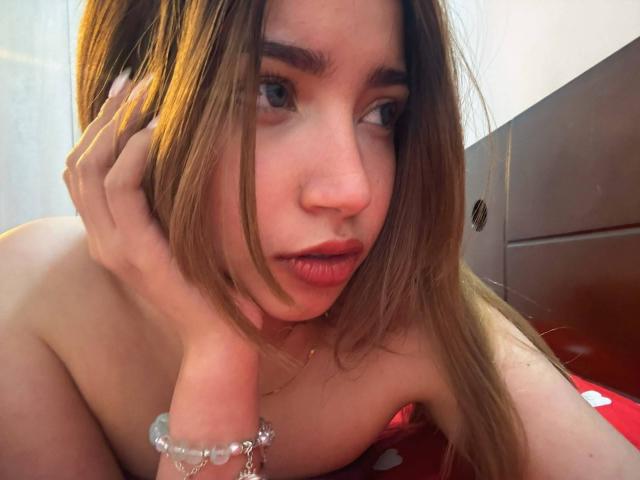 MirandaJones69 with long brown hair, blue eyes, and pink lips, looking at camera, hand on cheek, wearing a bracelet, topless, red bedsheet with white dots, wooden headboard.