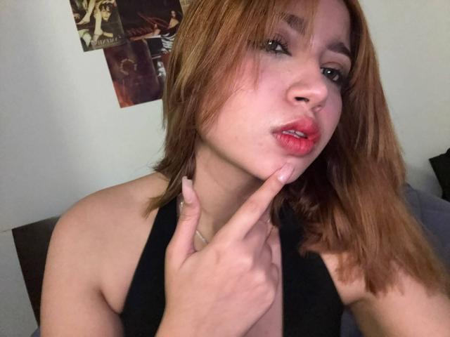MirandaJones69 with light brown hair, pouty red lips, and a finger touching her cheek, wearing a black halter top, showing her tits slightly. She's looking at the camera.