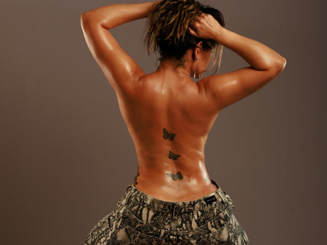 Topless JeniElfie with shiny, oiled skin, hands in messy brown hair, butterfly tattoos on back, wearing snake-patterned pants, arms raised, showing back and lower butt.