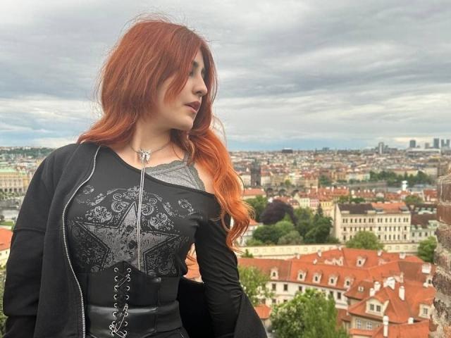 AssiaSou has red hair, black dress with intricate patterns, corset laces, and black cardigan. She's standing with her ass facing slightly away, looking to the side.