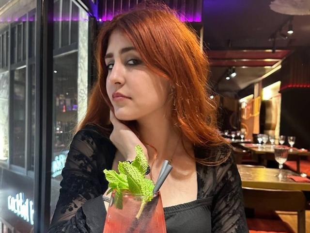 AssiaSou with red hair, black lace top, holding a drink with green leaves, looking to the side with a slight pout, in a dimly lit bar.