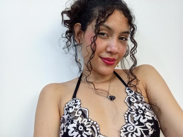 ValeryPoleth with curly brown hair, pink lipstick, nose ring, black lace bra, small tits, light brown skin, smiling, necklace with black heart, white background.