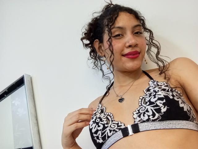 ValeryPoleth with curly black hair, wearing a black lace bra with white floral patterns, touching her bra, red lipstick, smiling, necklace with heart pendant, medium tits.
