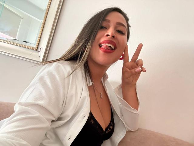 LiaDiane, com cabelos castanhos longos, batom vermelho, camisa branca aberta revelando um sutiã de renda preta, língua de fora, fazendo um sinal de paz, exibindo seus seios e um rosto bonito..