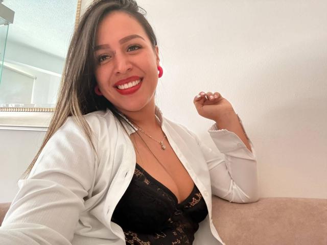 LiaDiane with brown hair, red lipstick, smiling, wearing a white shirt open to show black lace bra, red earrings, and necklace, holding up her right hand.