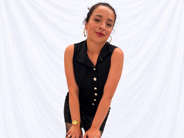 StangelarRivera, con el cabello oscuro recogido en un moño, vestida con una camisa negra sin mangas y con botones, con aretes de oro y un reloj de oro, inclinada hacia adelante, sonriendo, mostrando senos pequeños..