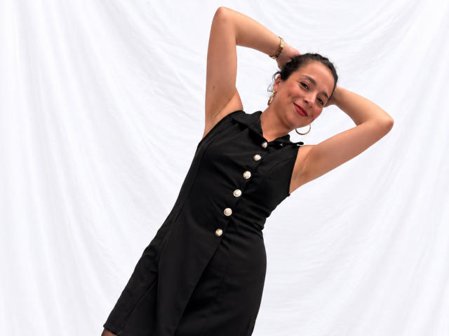 StangelarRivera, con los brazos levantados, vestido negro sin mangas, botones blancos, aros grandes, cabello oscuro, lápiz labial rojo, sonriendo, de pie frente a un fondo blanco con telas..
