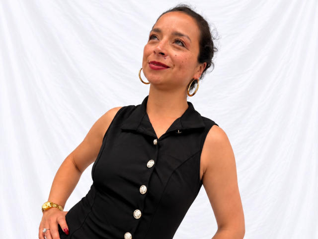 StangelarRivera with curly dark hair, wearing a black sleeveless button-up shirt, gold hoop earrings, red lipstick, and a gold bracelet, smiling confidently, showing her cute ass.