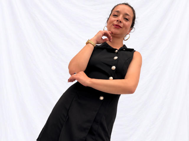 StangelarRivera, con cabello oscuro, vestida con un vestido negro abotonado, un brazalete dorado, aros grandes y una sonrisa, de pie frente a un fondo blanco, con la mano en la barbilla..