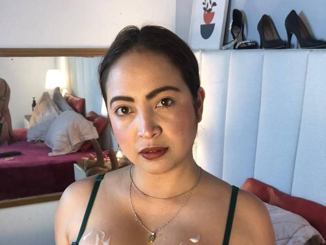 AlanaWest has medium brown skin, dark brown eyes, and brown hair in a low ponytail. She's wearing a green top, showing her tits, with a necklace and dark lipstick.