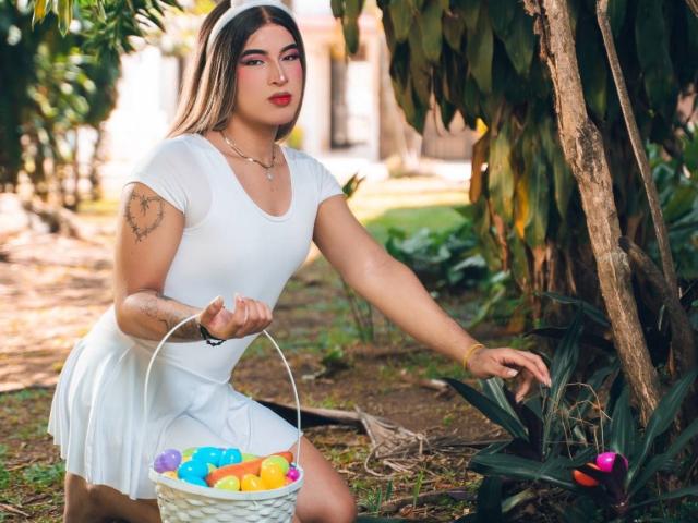 CamiRouxX mit gebräunter Haut, langen, dunklen Haaren, rotem Lippenstift, weißem Kleid, hält einen Osterkorb mit bunten Eiern, hat Tattoos am Arm, kniet im Garten und hat die rechte Hand auf einer Pflanze..