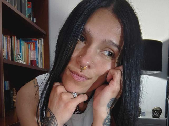 Amelie Blake, met lang zwart haar, een neusring en tatoeages, leunt met haar kin op haar handen, draagt een witte blouse en kijkt met een subtiele glimlach naar de camera..