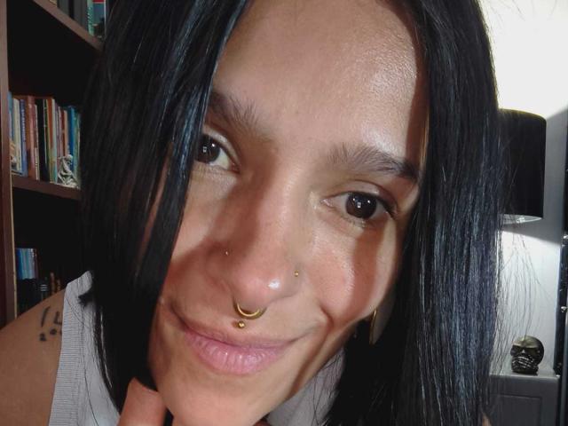 Amelie Blake heeft zwart haar dat tot op haar schouders reikt, bruine ogen, een neusring, een septumring en een kleine tatoeage op haar rechter schouder. Ze glimlacht en heeft haar hand in de buurt van haar kin..