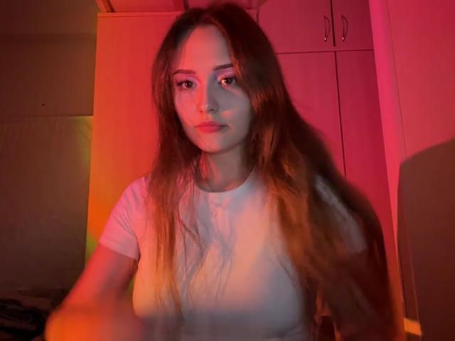 MinhMajic with long brown hair, white tee, and red-orange lighting, showing subtle tits, serious expression, in a dimly lit room.