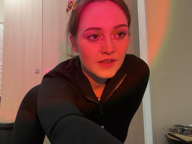 MinhMajic con il viso leggermente rosato, un felponcino nero con zip e i capelli raccolti in una clip colorata, è in avanti, guarda la telecamera con le labbra leggermente dischiuse, occhi semitrasparenti e riflessi rosa..