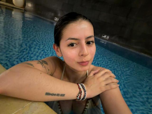 IsabellaJames with wet black hair, small tits, tattoo on right arm, bracelet on wrist, in blue-tiled pool, looking at camera, arms resting on pool edge.