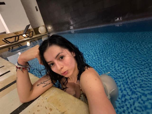 IsabellaJames with wet black hair, small tits, and a tattoo on her right arm, leaning on a pool edge, looking at the camera, wearing a white bikini.