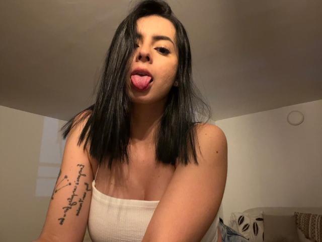 IsabellaJames with black hair, sticking out her tongue, wearing a white tube top, showing a tattoo on her right arm, in a dimly lit room.