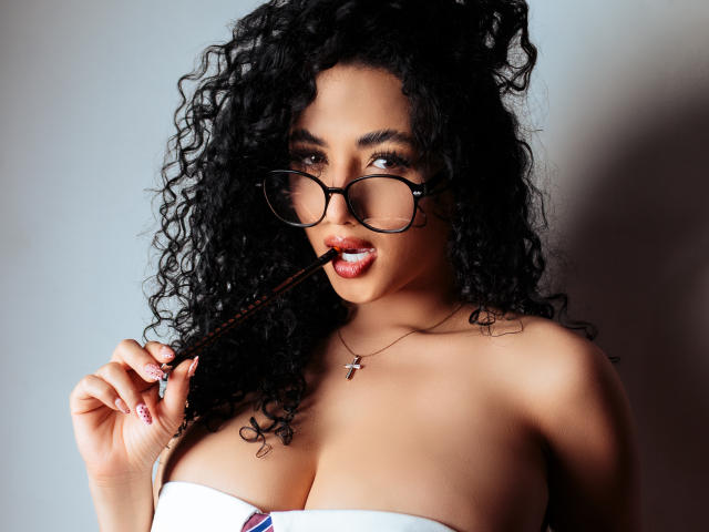 AuroraFerretti with curly black hair, wearing glasses, biting a brown stick, has large boobs, white strapless top, and a cross necklace, looking seductively at the camera.