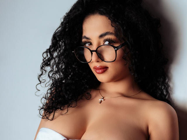 AuroraFerretti with curly black hair, wearing glasses, and a necklace, has her tits exposed, looking up with red lips, against a white background.