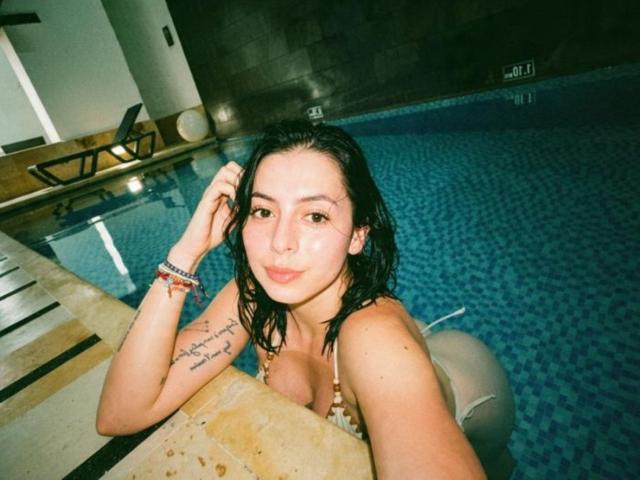 IsabellaJames with wet black hair, small tits, tattoo on left arm, in a blue-tiled pool, wearing a bikini, smiling, hand in hair, bracelet on wrist, indoor pool.