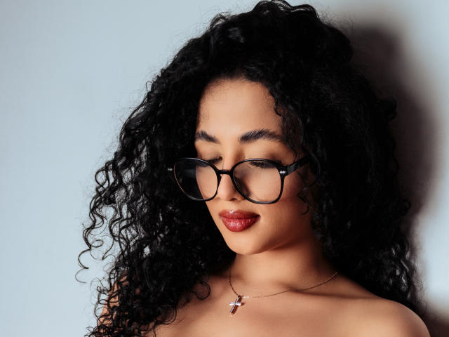 AuroraFerretti with curly black hair, wearing black glasses and red lipstick, has her eyes closed and a necklace with a red pendant, showing her bare shoulders.