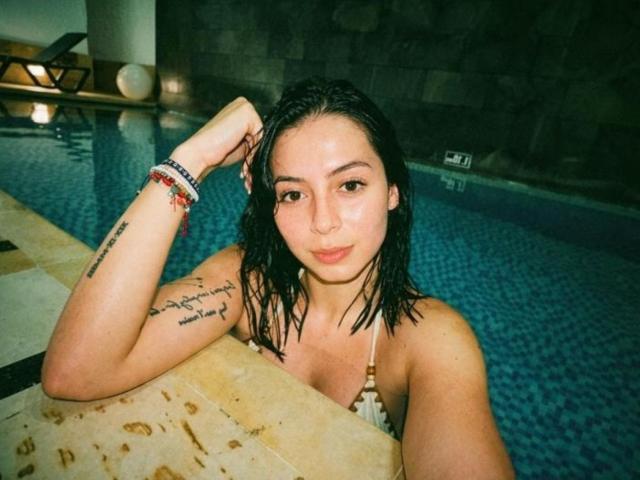 IsabellaJames has wet black hair, small tits, tattoo on left arm, wearing a patterned bikini, and red bead bracelet, leaning on pool edge, looking at camera.